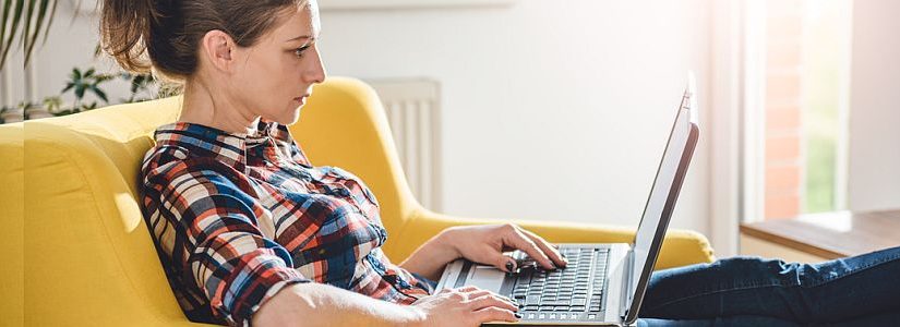 Heimarbeit - Frau arbeitet von zu Hause aus mit Laptop auf gelbem Sofa.